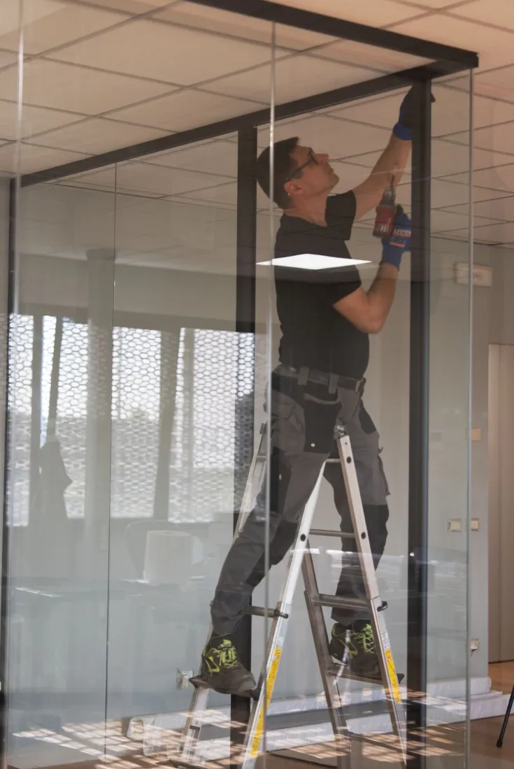 Installatore su scala che fissa con il trapano la struttura metallica nera di una parete divisoria in vetro